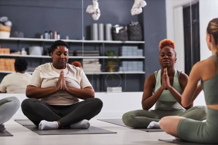 Téléchargez les photos : Femmes afro-américaines assis en position de lotus et méditant avec un instructeur pendant la formation - en image libre de droit