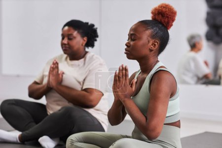 Téléchargez les photos : Afro-Américaine jeune femme assise en position de lotus et méditant en cours de yoga - en image libre de droit