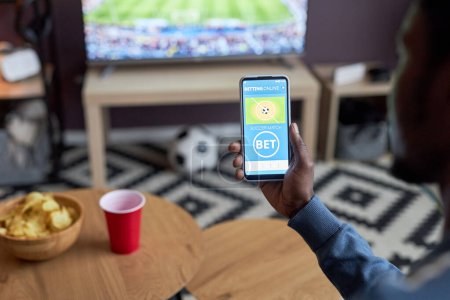 Téléchargez les photos : Gros plan du smartphone à main mâle avec paris sportifs en ligne à l'écran tout en regardant le match de football à la maison, espace de copie - en image libre de droit