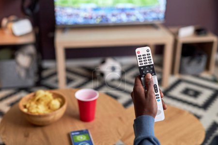 Foto de Primer plano del hombre viendo el partido de fútbol en casa con el foco en la mano masculina que sostiene la TV a distancia, espacio de copia - Imagen libre de derechos