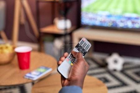 Téléchargez les photos : Gros plan du jeune homme noir regardant le match de football à la maison avec l'accent sur la main masculine tenant télécommande TV, espace de copie - en image libre de droit