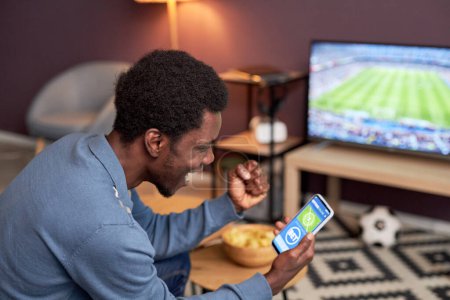 Téléchargez les photos : Portrait latéral de l'homme noir tenant smartphone avec l'application de paris sportifs en ligne et acclamant pour l'équipe de football - en image libre de droit