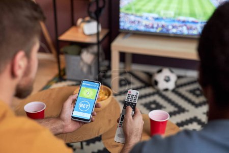 Foto de Primer plano de dos hombres viendo el partido de fútbol en la televisión y sosteniendo el teléfono inteligente con apuestas deportivas en línea - Imagen libre de derechos