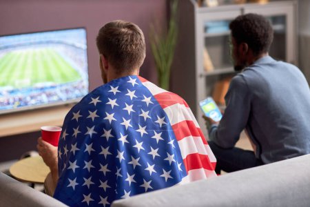 Téléchargez les photos : Vue arrière de deux hommes regardant un match de football à la télévision et portant le drapeau américain - en image libre de droit