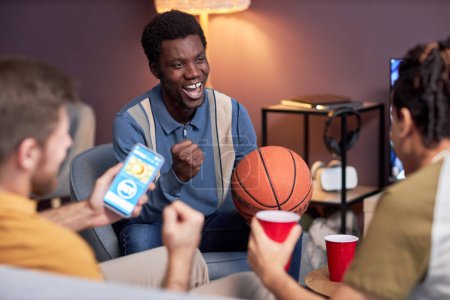 Foto de Retrato del hombre negro emocional animando para el partido de baloncesto en casa con amigos y sosteniendo la pelota - Imagen libre de derechos
