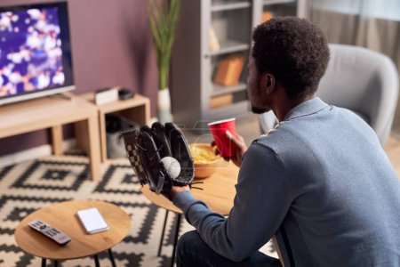Téléchargez les photos : Vue latérale du jeune homme noir portant un gant de baseball tout en regardant un match de sport à la maison, espace de copie - en image libre de droit
