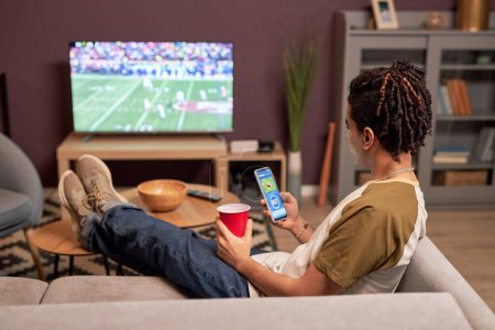 Téléchargez les photos : Vue arrière au jeune homme regardant le match de football à la maison et se détendre sur le canapé avec l'application de paris en ligne - en image libre de droit