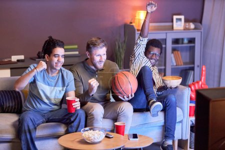 Foto de Diverso grupo de aficionados a los deportes emocionales viendo el partido de baloncesto en casa y animando iluminado por la luz azul - Imagen libre de derechos
