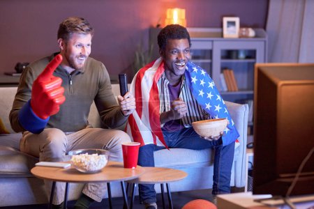 Téléchargez les photos : Portrait complet de deux fans de sport regardant la télévision en lumière bleue et portant le drapeau des États-Unis - en image libre de droit