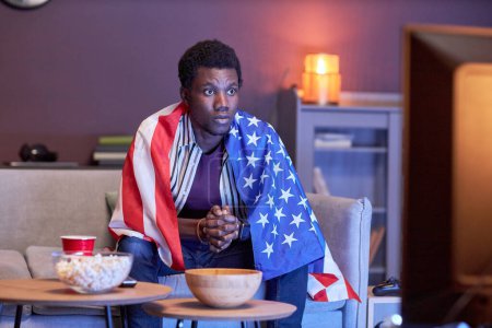 Téléchargez les photos : Portrait de jeune homme noir comme fan de sport regardant match à la télévision à la maison et portant le drapeau des États-Unis - en image libre de droit
