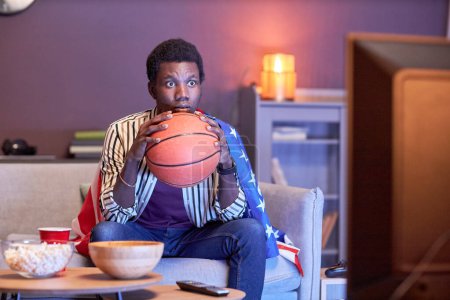 Téléchargez les photos : Portrait de jeune homme noir comme fan de sport regardant le match de basket-ball à la télévision à la maison et portant le drapeau des États-Unis en lumière bleue - en image libre de droit
