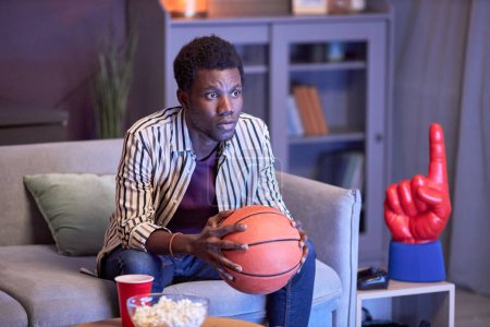 Téléchargez les photos : Portrait d'un jeune homme noir regardant un match de basket à la télé à la maison et tenant le ballon à la lumière bleue - en image libre de droit
