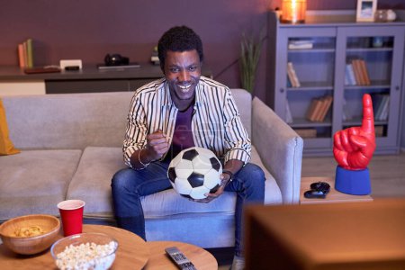Téléchargez les photos : Portrait de jeune homme noir regardant le match de football à la télévision à la maison et tenant le ballon dans la lumière bleue - en image libre de droit