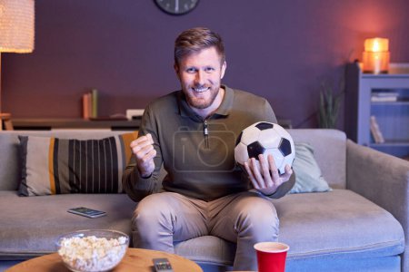 Foto de Retrato del hombre emocional viendo partidos deportivos en la televisión en casa y sosteniendo la pelota en luz azul, espacio de copia - Imagen libre de derechos