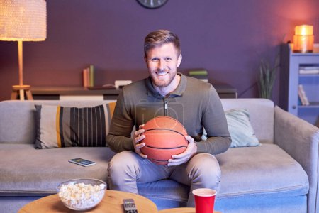 Téléchargez les photos : Portrait de l'homme émotionnel regardant match de sport à la télévision à la maison et tenant ballon de basket-ball dans la lumière bleue, espace de copie - en image libre de droit
