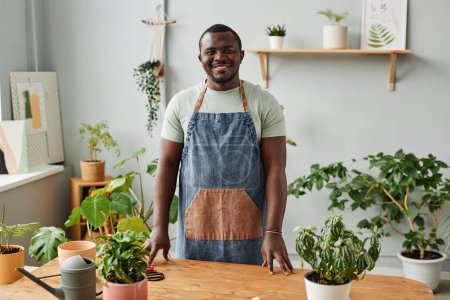 Téléchargez les photos : Portrait taille haute de l'homme noir adulte posant avec verdure luxuriante à l'intérieur et regardant la caméra - en image libre de droit