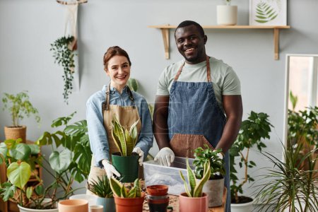 Téléchargez les photos : Portrait taille haute de deux jardiniers souriants regardant la caméra à l'intérieur tout en posant avec des plantes vertes luxuriantes - en image libre de droit