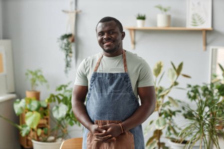 Téléchargez les photos : Portrait taille haute de l'homme noir souriant comme jardinier portant tablier posant à l'intérieur regardant la caméra avec des plantes vertes - en image libre de droit