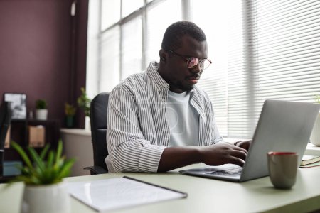 Photo for Portrait of focused black man using laptop while working at desk in office wearing casual clothes - Royalty Free Image