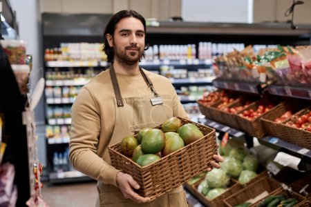 Photo for Waist up portrait of young man working in supermarket and carrying box with fresh exotic fruits - Royalty Free Image