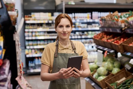 Foto de Retrato de cintura hacia arriba de una mujer joven que trabaja en el supermercado y sonríe a la cámara sosteniendo la tableta - Imagen libre de derechos