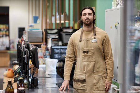 Photo for Portrait of bearded young man working in supermarket and looking at camera standing by cashiers desk, copy space - Royalty Free Image