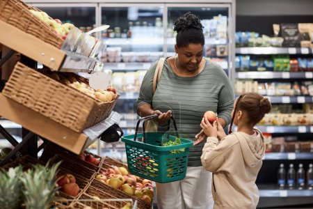 Photo for Portrait of real black woman shopping in supermarket with little daughter and buying fresh vegetables - Royalty Free Image