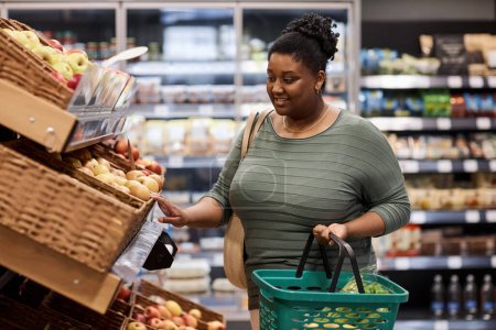 Téléchargez les photos : Portrait à la taille d'une femme noire souriante faisant ses courses dans un supermarché et achetant des fruits frais - en image libre de droit