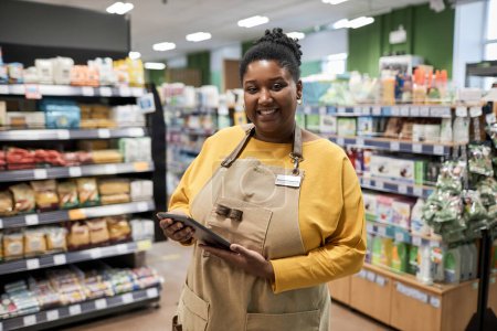 Foto de Retrato de cintura hacia arriba de una mujer negra sonriente trabajando en el supermercado y mirando alegremente a la cámara - Imagen libre de derechos