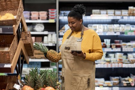 Photo for Side view portrait of smiling black woman enjoying work in supermarket and inspecting exotic fruits - Royalty Free Image