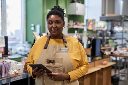 Foto de Cintura hacia arriba retrato de mujer negra real disfrutando del trabajo en el supermercado y mirando a la cámara - Imagen libre de derechos