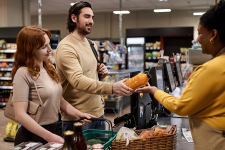 Téléchargez les photos : Portrait latéral de jeune couple debout près du bureau des caissiers dans un supermarché et achetant des produits d'épicerie - en image libre de droit