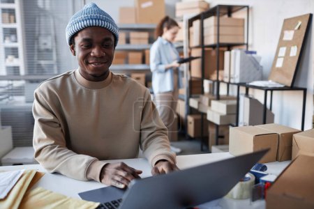 Téléchargez les photos : Jeune homme afro-américain souriant en tenue décontractée assis par le lieu de travail devant un ordinateur portable dans la salle de stockage et de prendre de nouvelles commandes en ligne - en image libre de droit