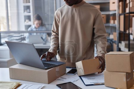 Photo for Cropped shot of black guy in sweatshirt checking numbers of packed parcels while standing by workplace in front of laptop in post office - Royalty Free Image