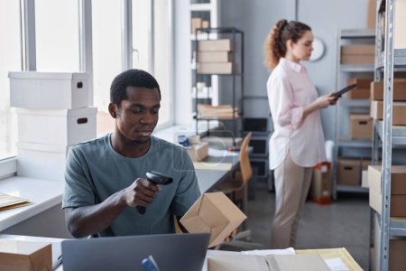 Téléchargez les photos : Jeune Afro-Américain assis par lieu de travail et numérisant le code sur une boîte en carton contenant des marchandises commandées par les consommateurs en ligne - en image libre de droit