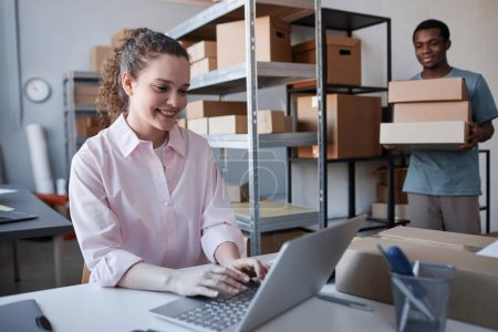 Photo for Happy young woman typing on laptop keyboard while African American man carrying stack of cardboard boxes containing ordered goods - Royalty Free Image