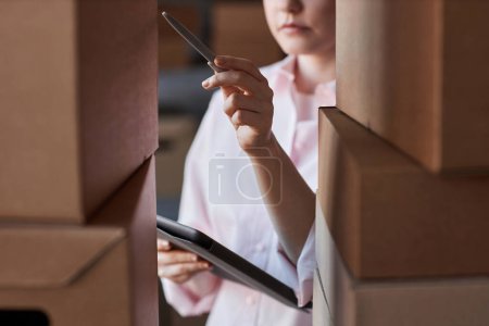 Téléchargez les photos : Gros plan de la jeune femme avec stylo et tablette pointant vers l'une des parcelles dans une pile de boîtes en carton avec des marchandises commandées pendant le travail - en image libre de droit