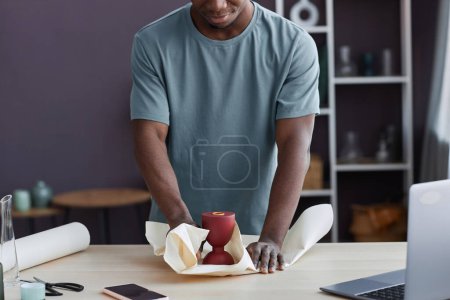 Téléchargez les photos : Plan recadré d'un jeune artisan enveloppant une petite tasse rouge faite à la main dans du papier tout en restant debout devant un bureau devant un ordinateur portable dans un atelier - en image libre de droit