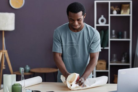 Téléchargez les photos : Jeune Afro-Américain propriétaire de magasin d'artisanat emballage ordre de client en ligne dans du papier avant de l'emballer dans une boîte et l'envoyer au récepteur - en image libre de droit