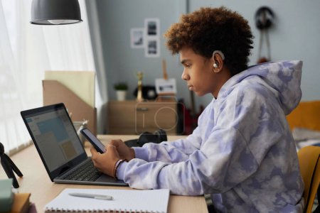 Téléchargez les photos : Vue latérale de l'adolescent afro-américain textos dans le smartphone tout en étant assis par le bureau en face de l'ordinateur portable et en choisissant la musique de la liste de lecture - en image libre de droit