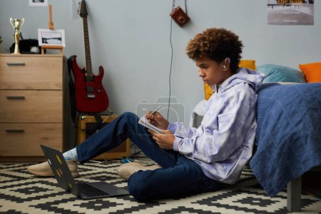Téléchargez les photos : Adolescent afro-américain dans les écouteurs prendre des notes dans le copybook pendant la conférence en ligne tout en étant assis devant un ordinateur portable sur le sol près de son lit - en image libre de droit