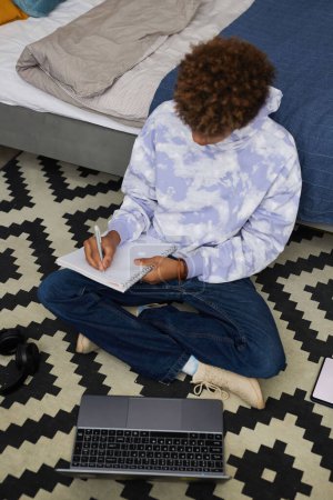 Foto de Alto ángulo de adolescente afroamericano haciendo notas en copybook mientras está sentado en el suelo y preparándose para el seminario escolar - Imagen libre de derechos