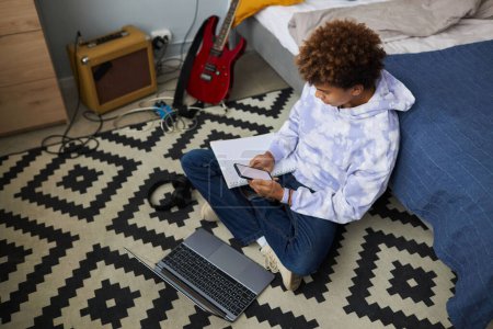 Colegial joven en sudadera con capucha y jeans clásicos usando el teléfono móvil mientras está sentado en el suelo frente a la computadora portátil y preparando la tarea