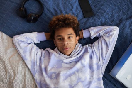 Téléchargez les photos : Jeune garçon serein en sweat à capuche élégant regardant la caméra tout en étant couché sur un lit double confortable bleu et relaxant après avoir fait des devoirs - en image libre de droit