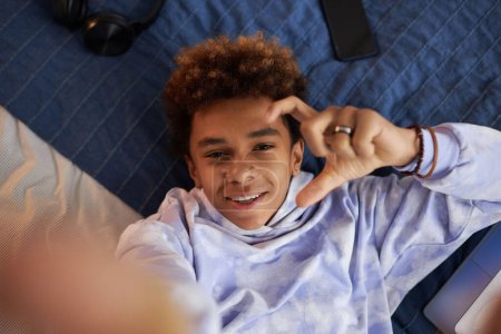 Téléchargez les photos : Focus sur le visage de joyeux adolescent afro-américain regardant la caméra avec le sourire tout en se relaxant sur le lit à loisir et en prenant selfie - en image libre de droit
