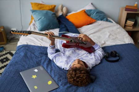 Téléchargez les photos : Reposé adolescent afro-américain jouant de la guitare électrique tout en se relaxant sur un lit double confortable après l'école et en profitant de loisirs - en image libre de droit