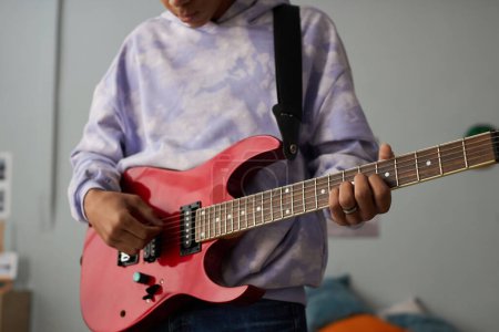 Téléchargez les photos : Gros plan d'un adolescent en sweat-shirt tenant une guitare électrique tout en se tenant devant la caméra et apprenant à jouer d'un instrument de musique à cordes - en image libre de droit