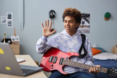 Téléchargez les photos : Adolescent heureux en tenue décontractée saluant son professeur de musique en agitant la main tout en étant assis sur le lit devant un ordinateur portable avant la leçon en ligne - en image libre de droit
