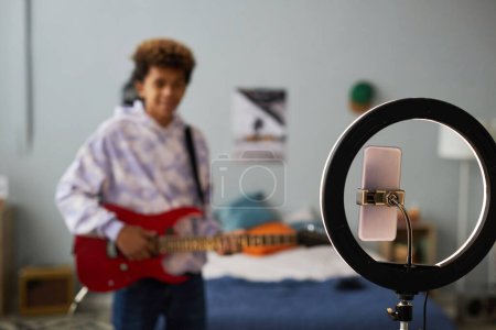 Téléchargez les photos : Focus sur cadre rond avec smartphone sur support lors de l'enregistrement vidéo contre un adolescent jouant de la guitare électrique dans sa chambre - en image libre de droit