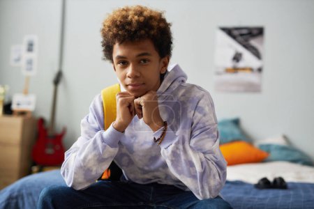 Photo for Cute serene teenage boy in casualwear keeping hands by chin while sitting on double bed in his bedroom at leisure and looking at camera - Royalty Free Image